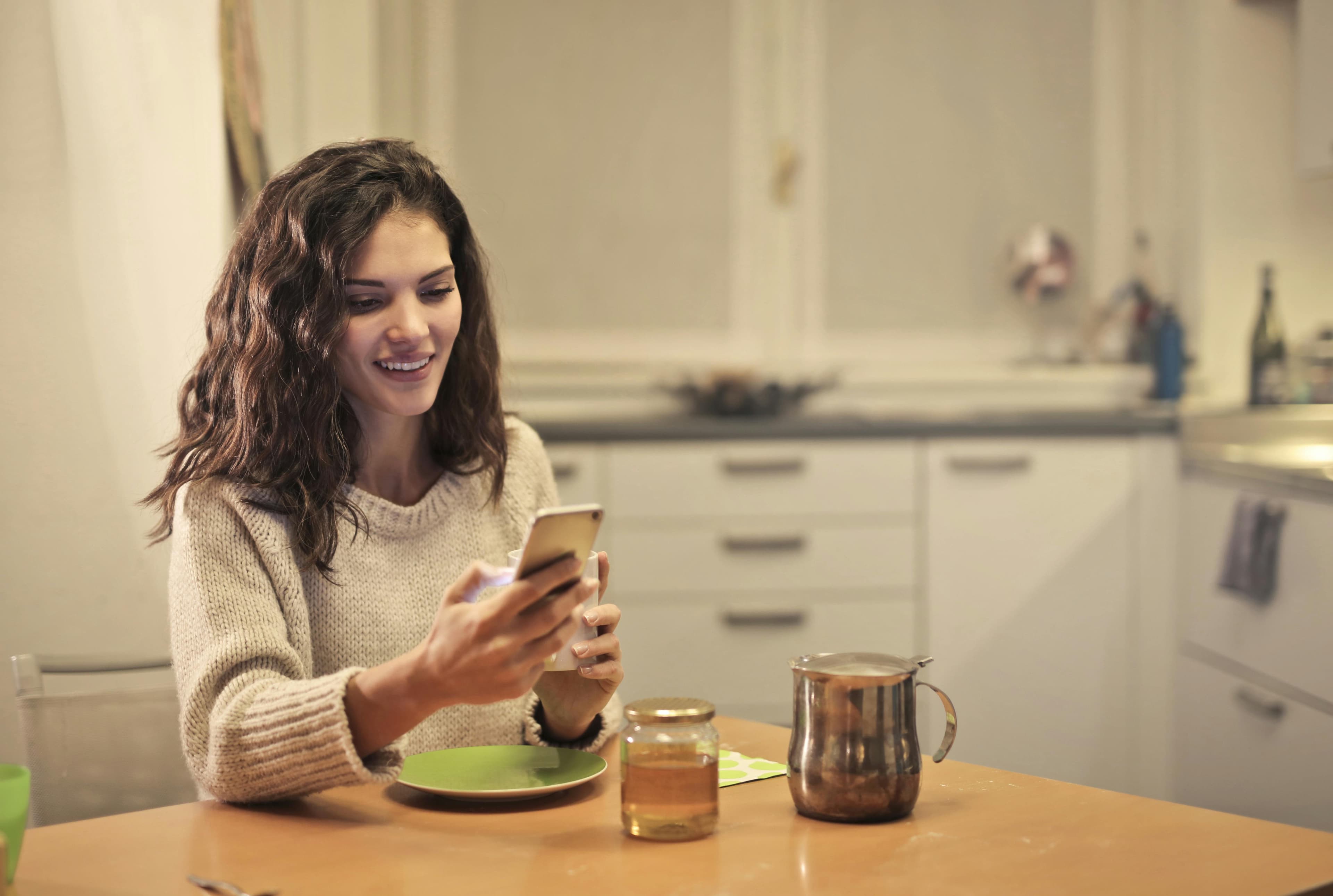 Woman at kitchen table smiling at phone with warm cup of tea